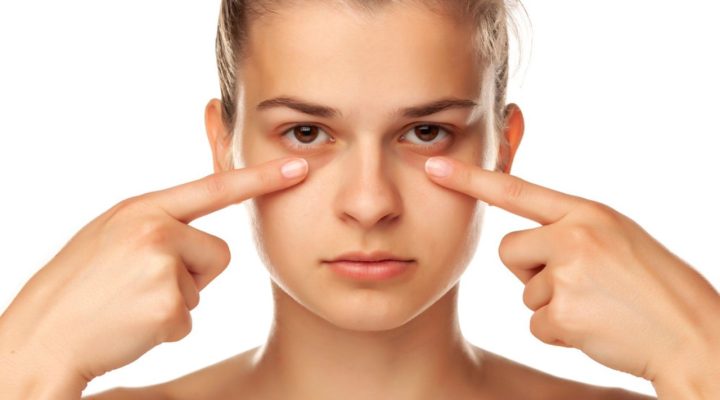 Jeune femme montrant ses cernes du bout des doigts, regard neutre, sur fond blanc.