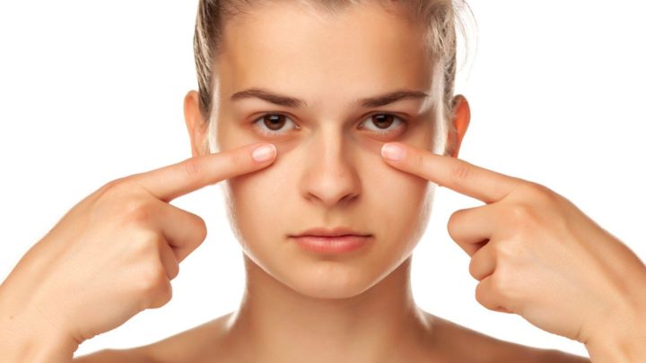 Jeune femme montrant ses cernes du bout des doigts, regard neutre, sur fond blanc.
