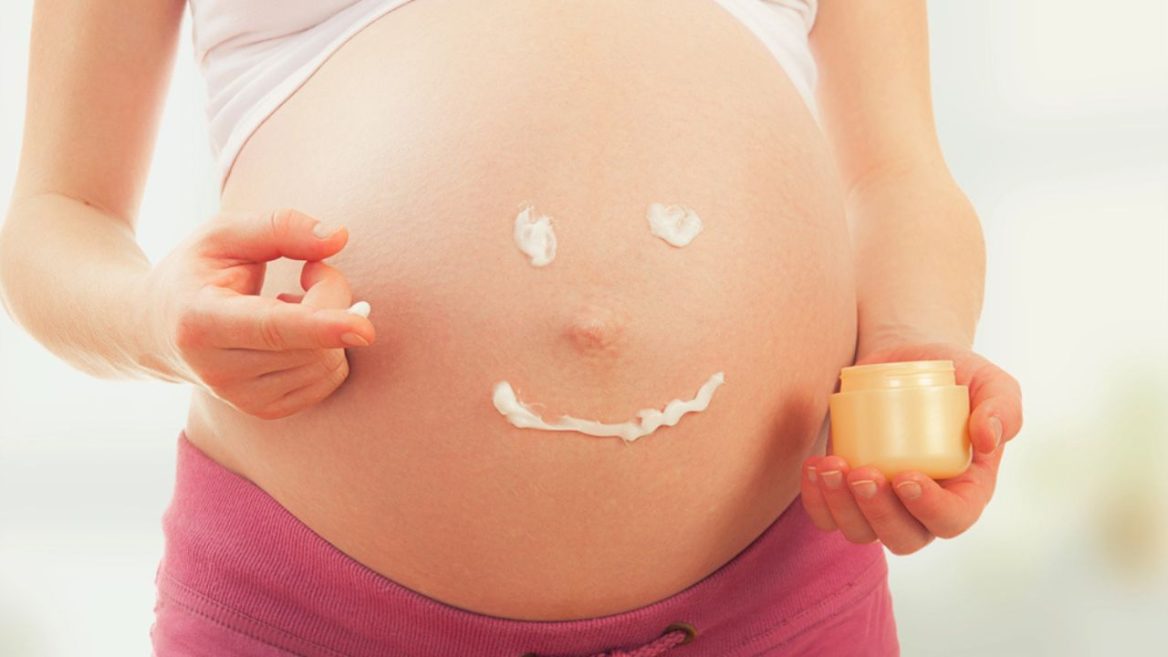 Femme enceinte appliquant une crème sur son ventre, avec un visage souriant dessiné en crème sur la peau, illustrant les soins esthétiques pendant la grossesse.