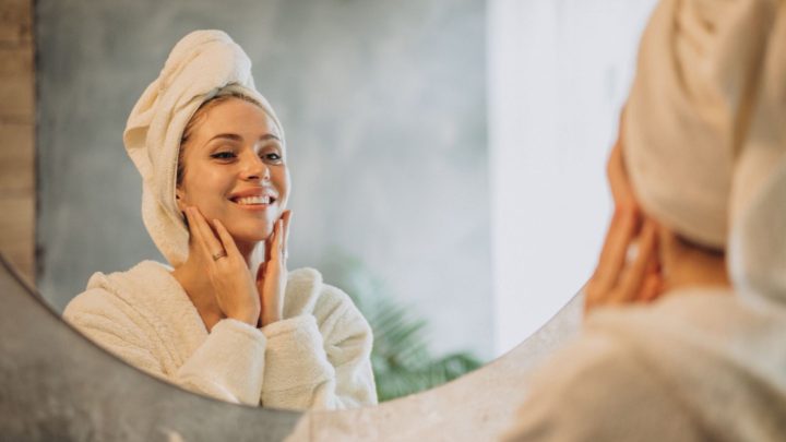 Femme souriante en peignoir devant un miroir, prenant soin de sa peau pour préserver l’équilibre du microbiote cutané